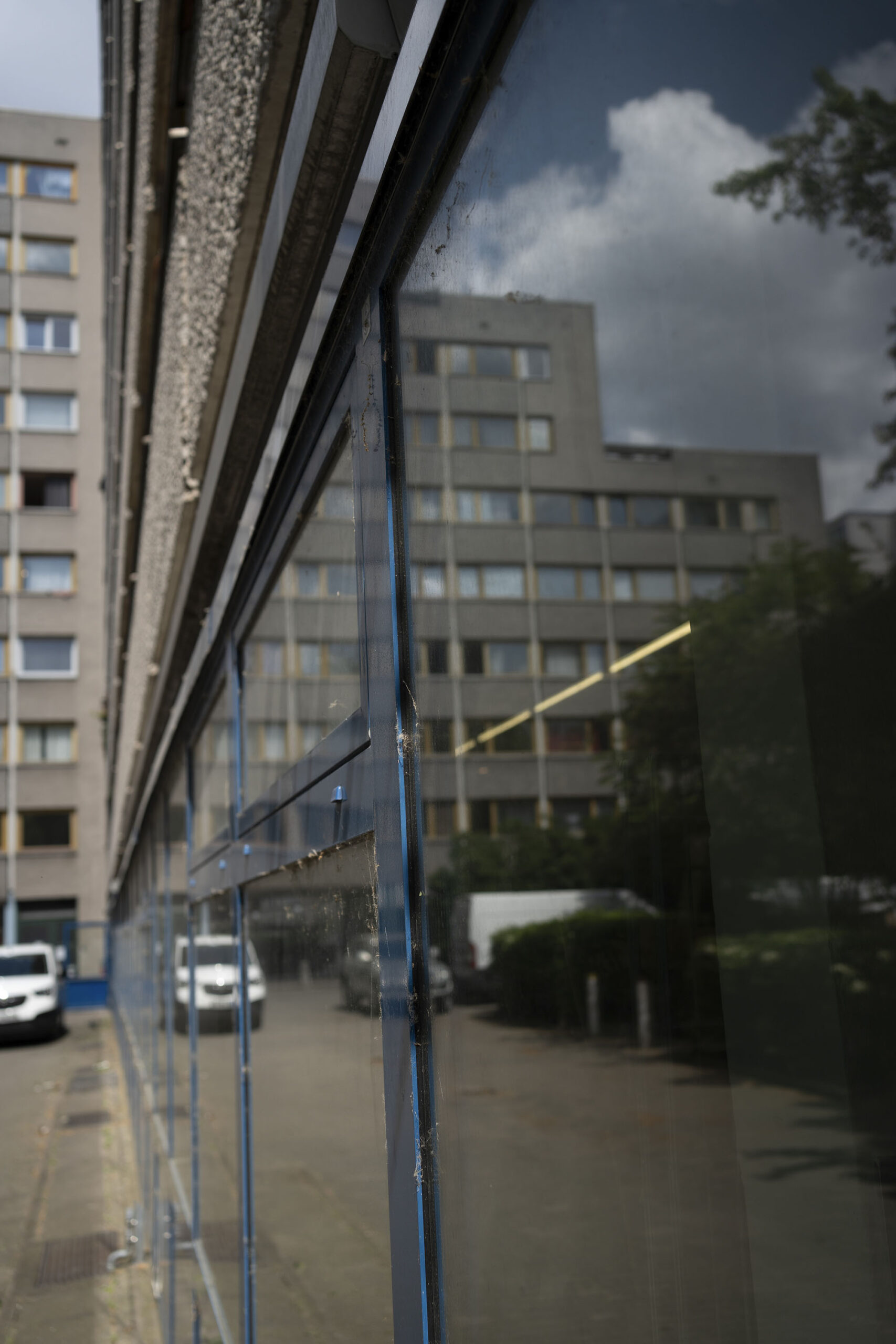 Eine Reihe von Wohnhäusern mit reflektierenden Glasplatten und einem geparkten weißen Auto im Hintergrund. Bewölkter Himmel und Baumlaub sind ebenfalls sichtbar. (KI generierte Beschreibung)