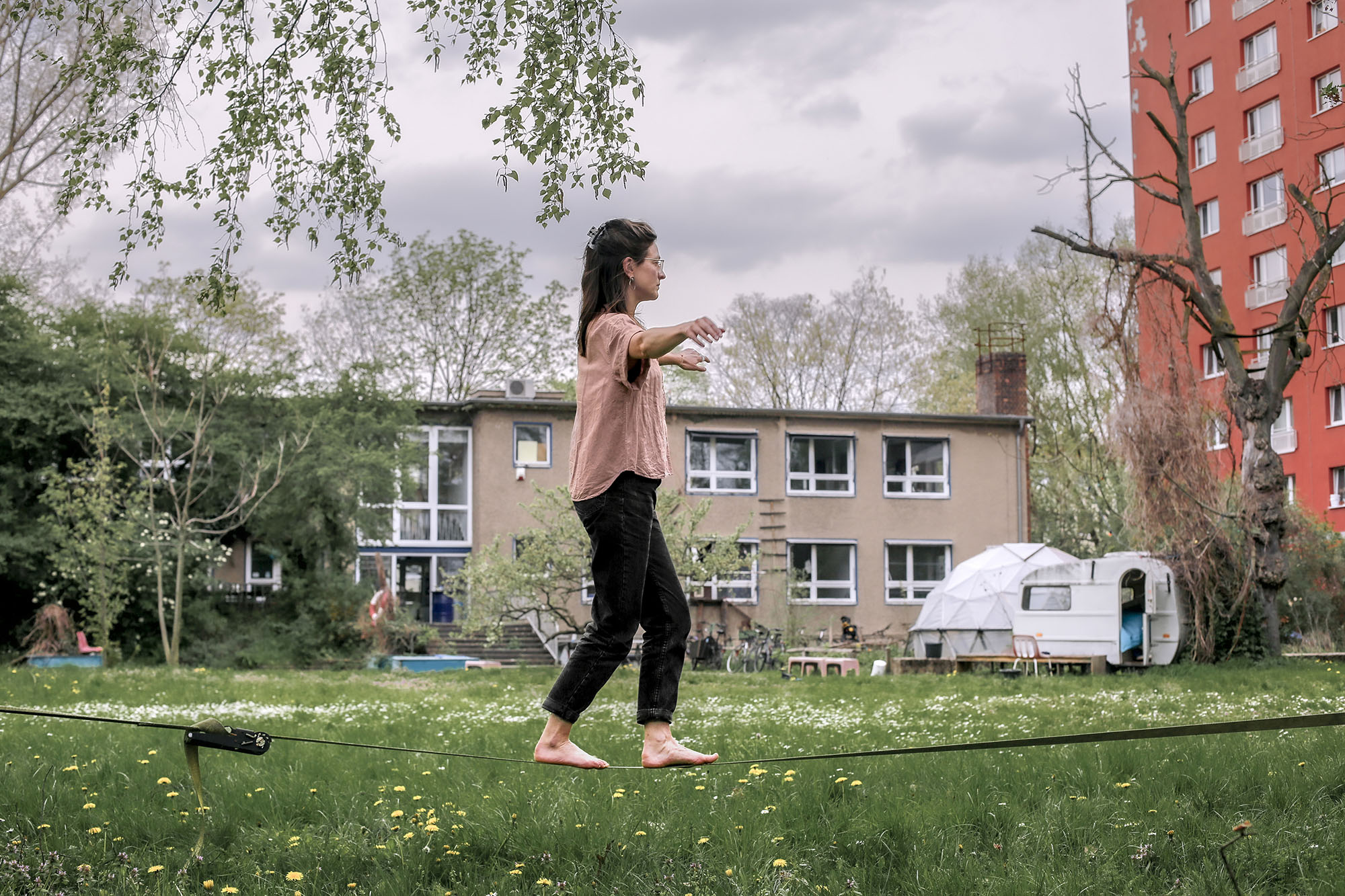 Person, die auf einer Slackline in einer Rasenfläche balanciert, mit Gebäuden und Bäumen im Hintergrund. (KI generierte Beschreibung)