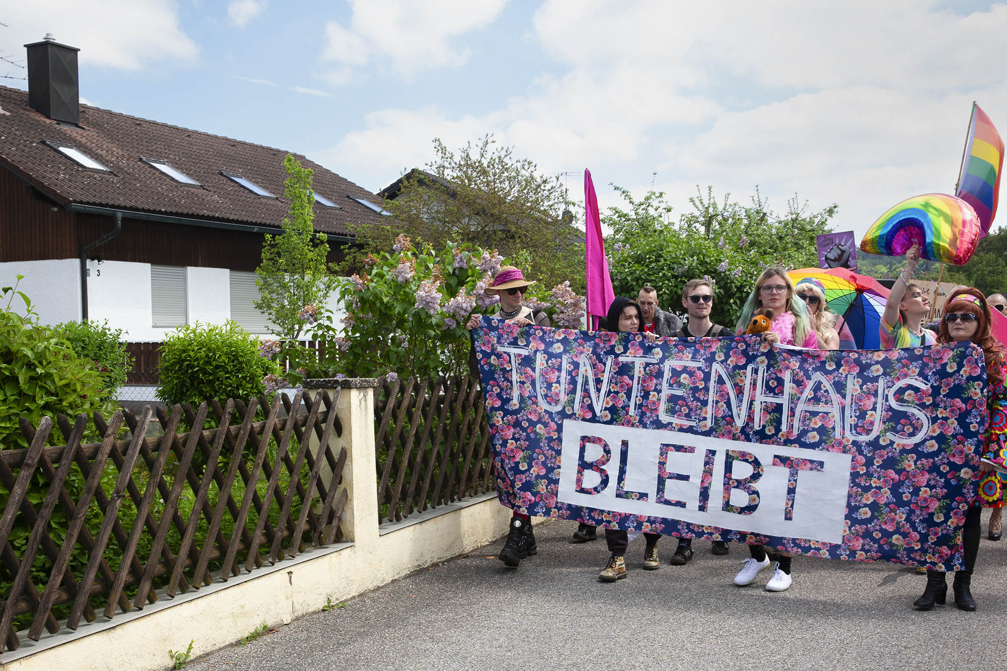 Eine Gruppe von Menschen in bunten Outfits hält während einer Protestkundgebung ein Banner mit der Aufschrift „Tuntenhaus bleibt“. Sie laufen eine Straße entlang, die an einem Zaun und Wohnhäusern vorbeiführt. (KI generierte Beschreibung)