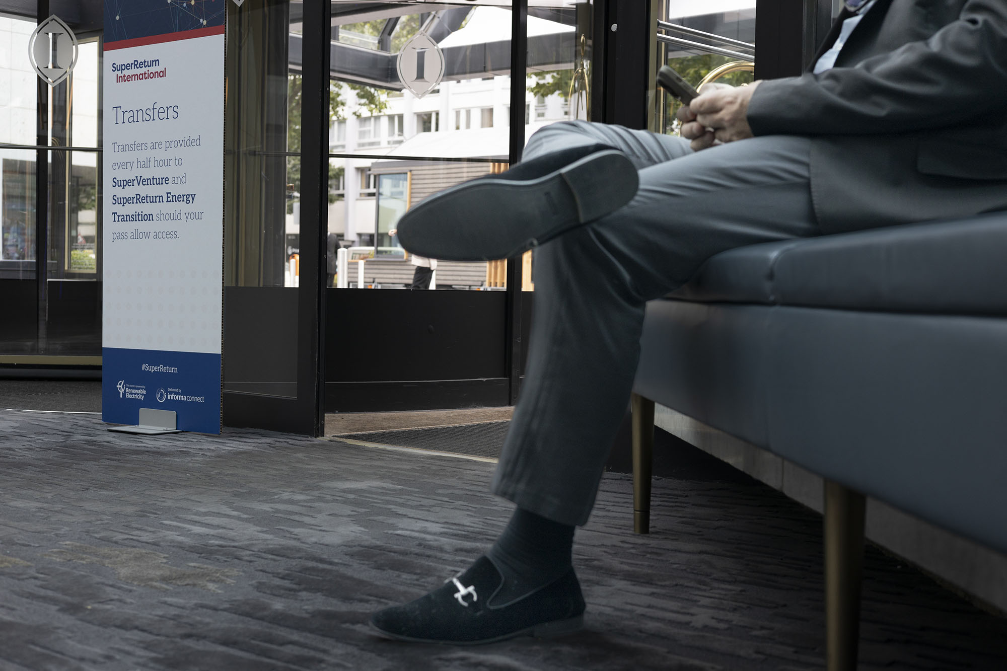 Person in Geschäftskleidung sitzt auf einer Bank und hält ein Telefon. Ein Schild im Hintergrund weist auf die Transferdienste von SuperReturn International hin. (KI generierte Beschreibung)