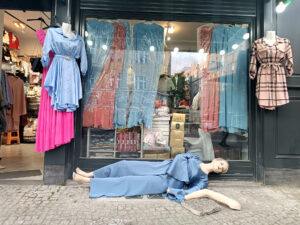 Eine Schaufensterpuppe im blauen Kleid liegt auf dem Bürgersteig vor einem Bekleidungsgeschäft, umgeben von hängenden Kleidern und einer Schaufensterauslage. (KI generierte Beschreibung)