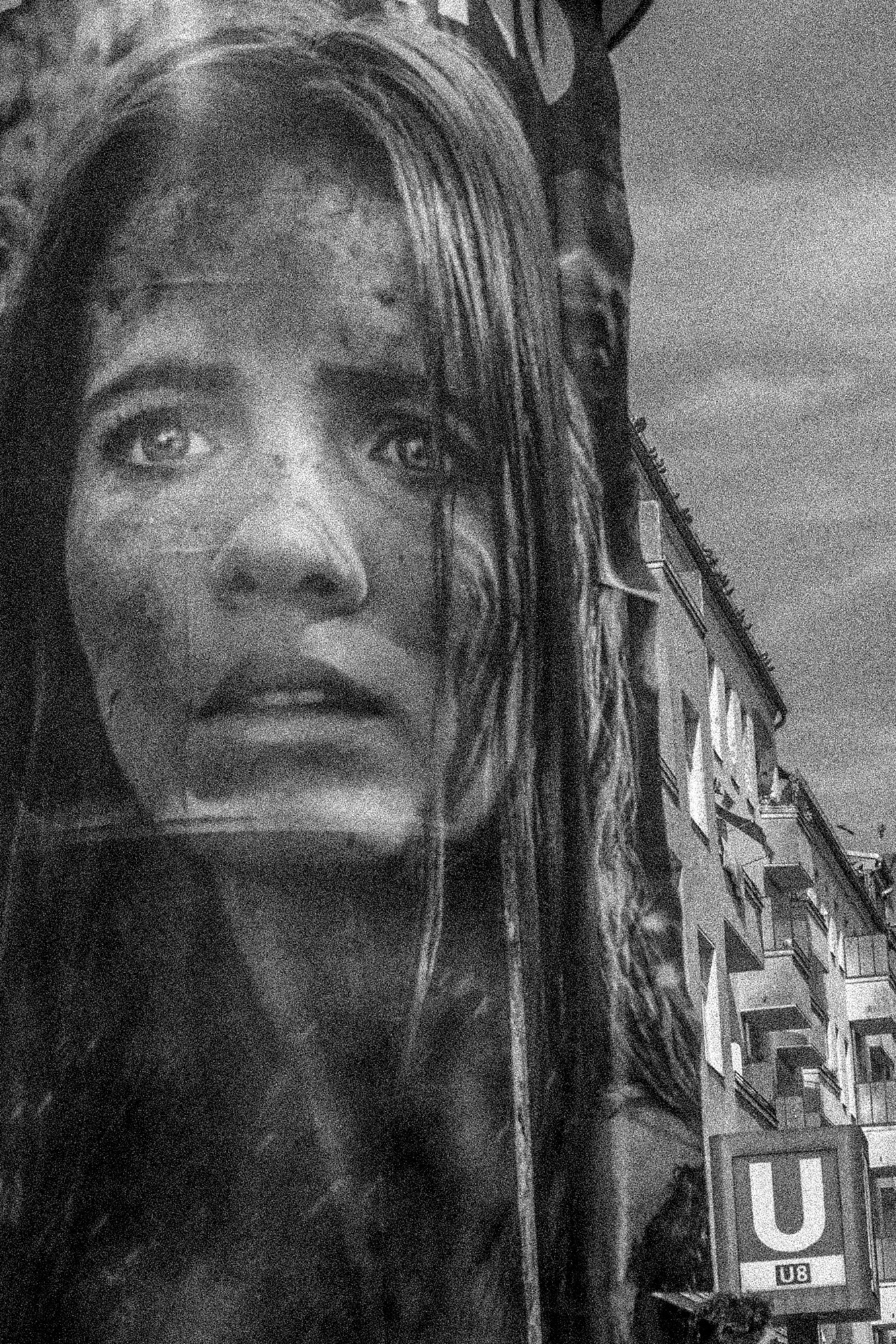 Ein Schwarzweißfoto eines großen Plakats, das eine Nahaufnahme eines Frauengesichts zeigt, teilweise verwittert. Im Hintergrund sind Gebäude und ein U-Bahn-Eingang mit einem „U8“-Schild zu sehen. (KI generierte Beschreibung)