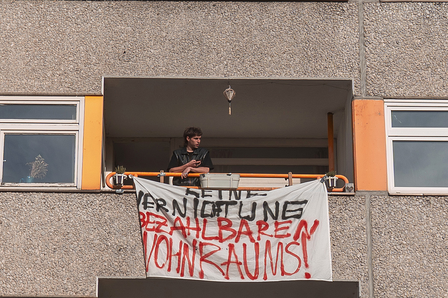Person steht auf einem Balkon mit einem Transparent mit der Aufschrift „Keine Verdrängung Bezahlbaren Wohnraums!“ auf der Außenseite eines Wohngebäudes. (KI generierte Beschreibung)
