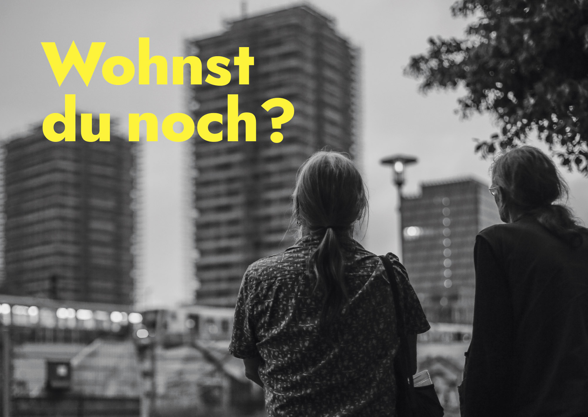 Zwei Menschen mit langen Haaren beobachten eine vorbeifahrende U-Bahn in einer städtischen Umgebung. Der Text lautet: „Wohnst du noch?“ (KI generierte Beschreibung)