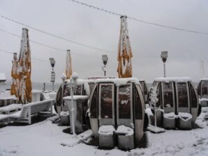 Schneebedeckter Sitzbereich im Freien mit runden Schirmen und hohen Sonnenschirmen. Lichterketten hängen darüber und der Boden ist unter einem bewölkten Himmel mit Schnee bedeckt. (KI generierte Beschreibung)