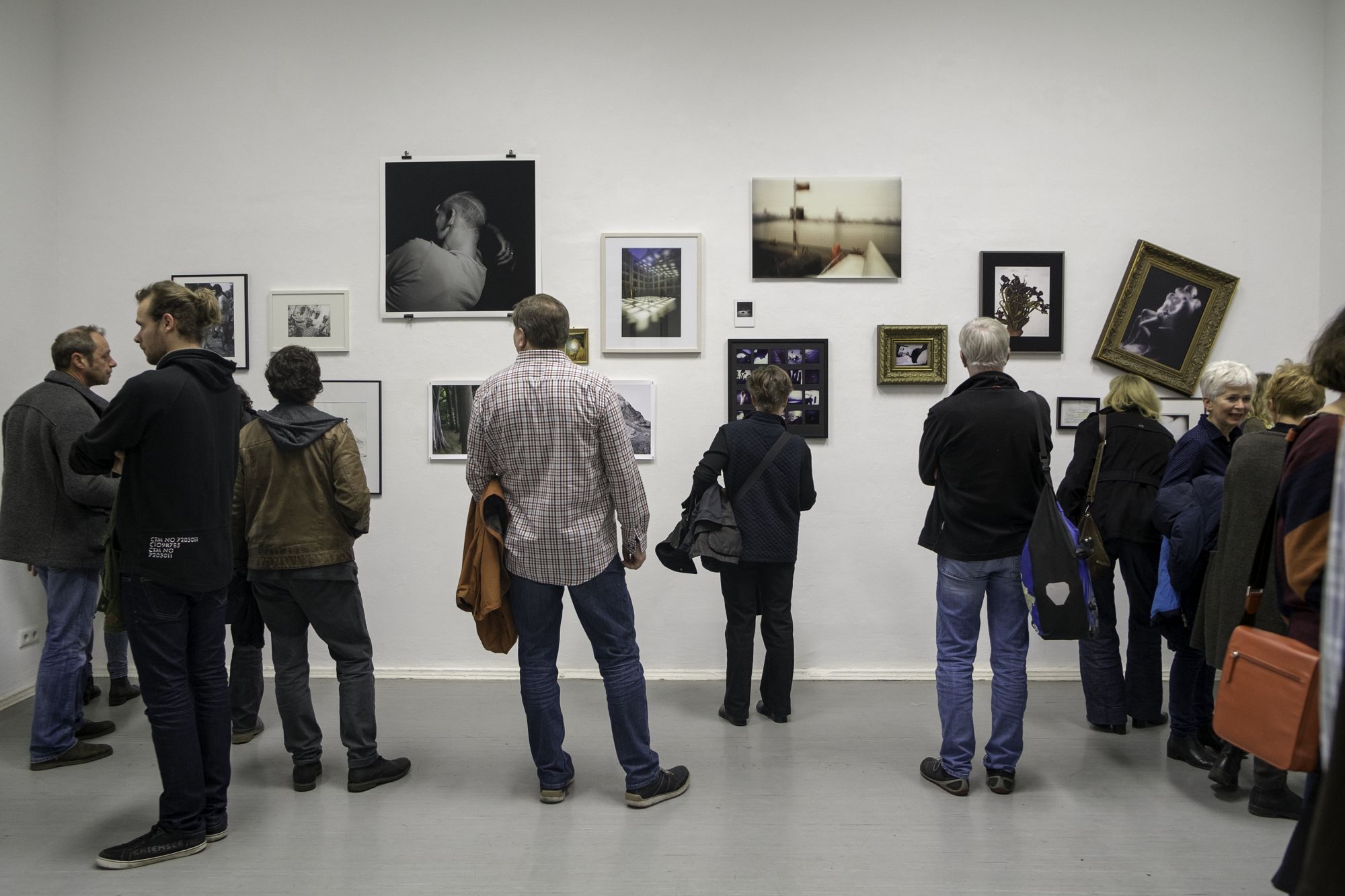 Menschen betrachten in einer Galerie verschiedene gerahmte Kunstwerke, einige schief, andere aufrecht. (KI generierte Beschreibung)