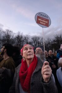 Person, die während einer Versammlung im Freien in einer Menschenmenge ein Schild hält und einen roten Schal und ein rosa Stirnband trägt. Im Hintergrund sind Bäume unter einem bewölkten Himmel zu sehen. (KI generierte Beschreibung)