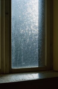 Milchglasfenster mit durchscheinendem Licht, das einen strukturierten, funkelnden Effekt auf dem Glas erzeugt. Ein hölzerner Fensterrahmen und eine Fensterbank sind sichtbar, wobei teilweise Licht auf die Fensterbank fällt. (KI generierte Beschreibung)