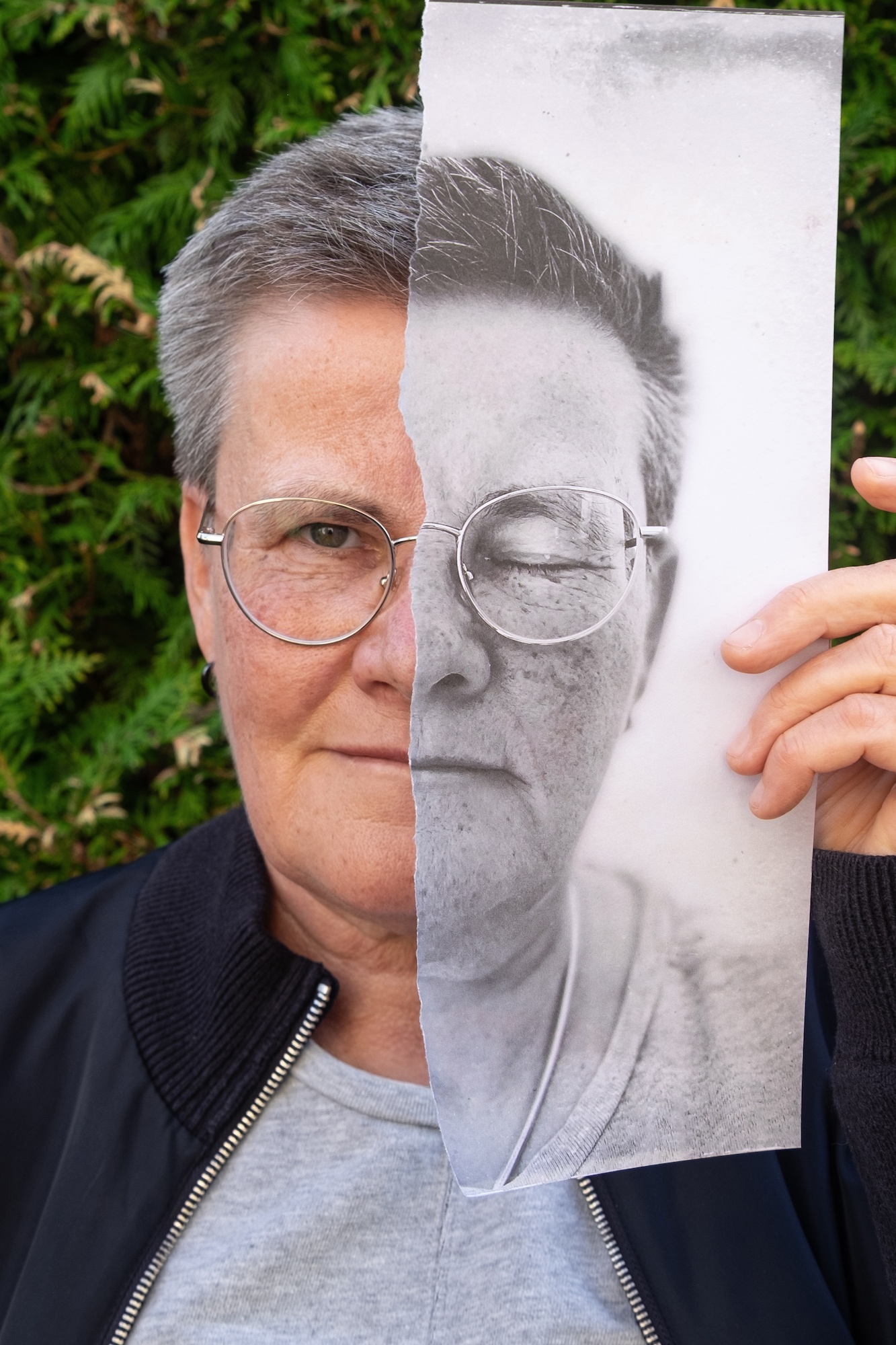 Eine ältere Person mit kurzen grauen Haaren und Brille hält ein zerrissenes Schwarz-Weiß-Foto ihres eigenen Gesichts in der Hand und richtet es über der Hälfte ihres echten Gesichts aus. Im Hintergrund ist grünes Laub zu sehen. (KI generierte Beschreibung)