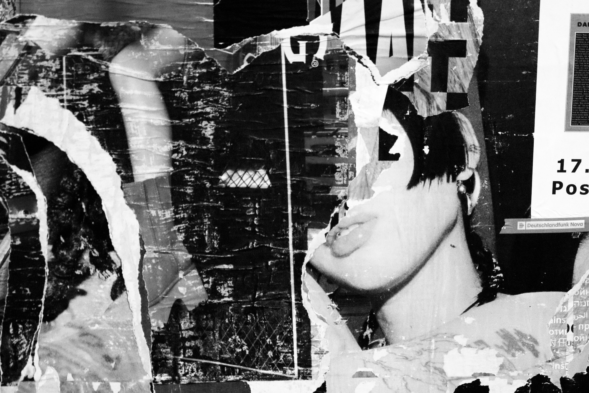 Ein Schwarz-Weiß-Foto einer Wand mit sich überlappenden, zerrissenen Plakaten, darunter ein teilweise sichtbares Frauengesicht mit kurzen Haaren und geschürzten Lippen. (KI generierte Beschreibung)