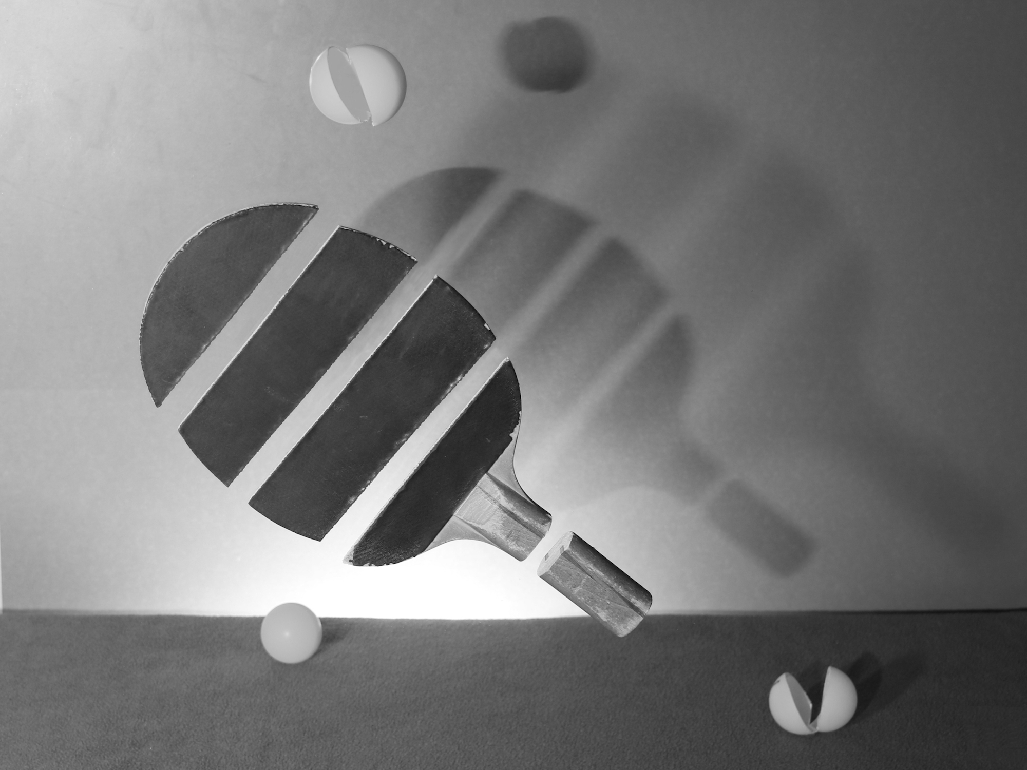 Ein Schwarz-Weiß-Foto von einem Tischtennisschläger mit drei Tischtennisbällen und deren Schattenwurf an der Wand dahinter. (KI generierte Beschreibung)