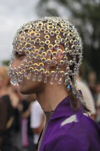 Eine Person, die eine Kopfbedeckung aus schillernden Ringen und tropfenförmigen Kristallen trägt, steht in einem lilafarbenen Outfit im Freien inmitten einer Menschenmenge. (KI generierte Beschreibung)