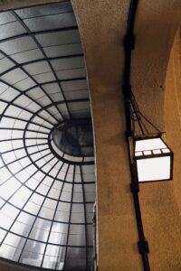 Eine Glaskuppel mit Gittermuster auf der linken Seite und eine beleuchtete quadratische Laterne, die von einem strukturierten Steinbogen auf der rechten Seite hängt. (KI generierte Beschreibung)