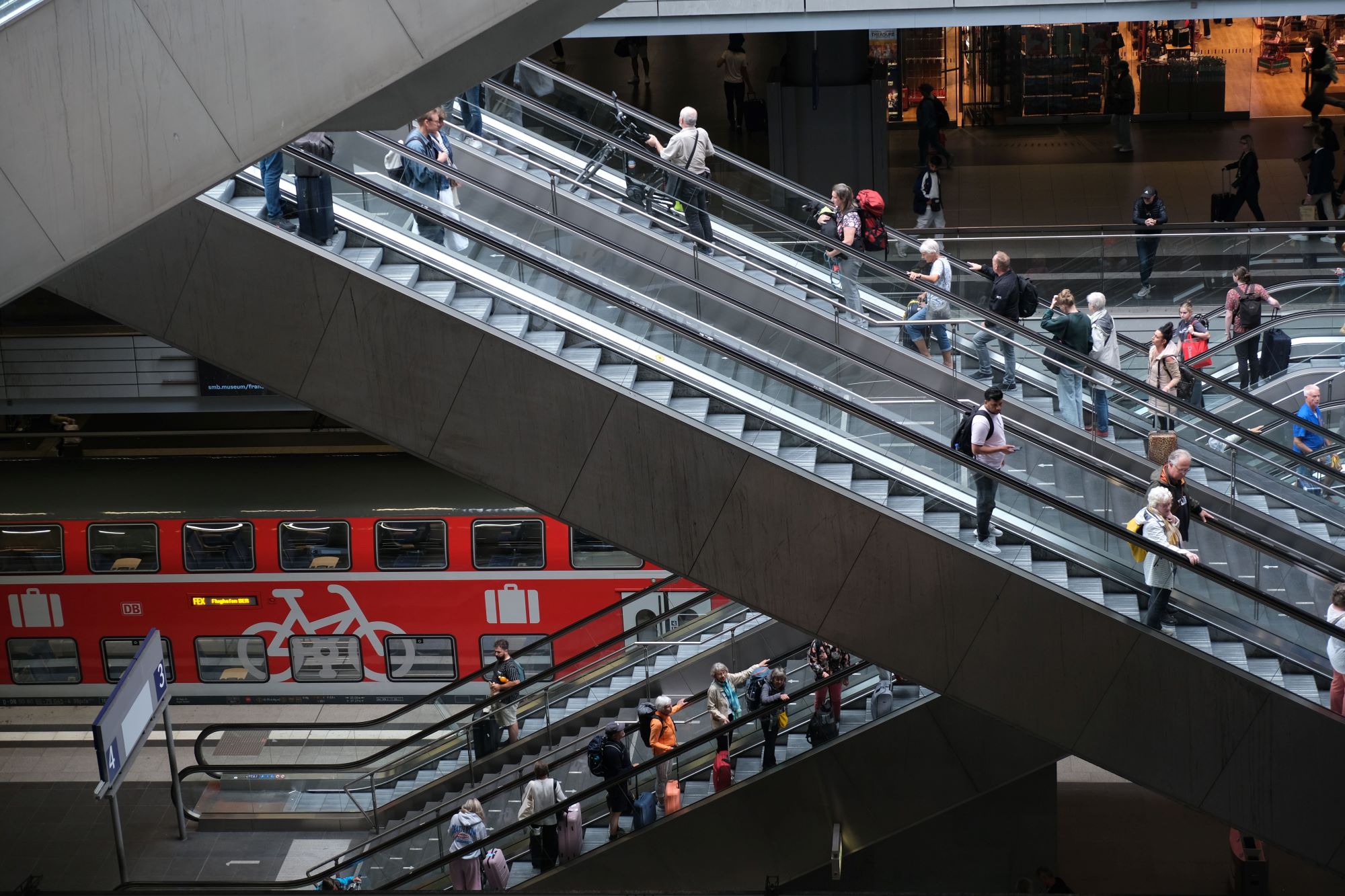 In einem modernen Bahnhof benutzen die Menschen mehrere Rolltreppen, wobei auf der unteren Ebene ein roter Zug zu sehen ist und die Fahrgäste zwischen den Etagen wechseln. (KI generierte Beschreibung)