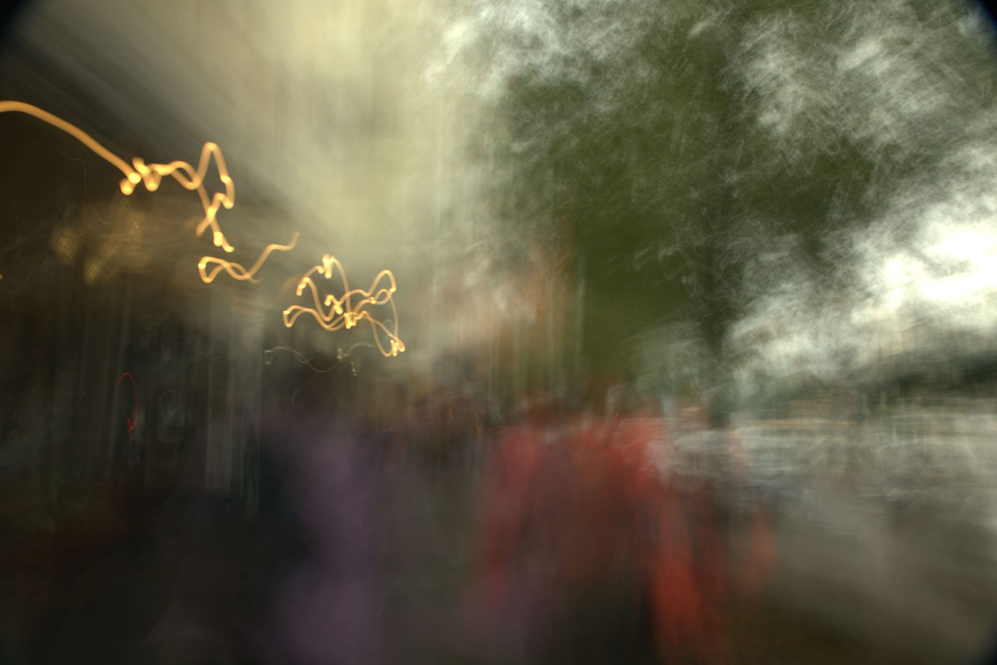 Eine verschwommene, abstrakte Szene im Freien mit gelben Lichtstreifen, undeutlichen Figuren und Bäumen im Hintergrund. (KI generierte Beschreibung)