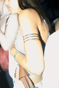 Eine Frau in einem weißen Spitzenoberteil mit einem braunen Gürtel und einer metallischen Armmanschette steht in einer Menschenmenge, teilweise von der Kamera abgewandt. (KI generierte Beschreibung)