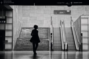 Eine Person geht auf eine Treppe und eine Rolltreppe in einer modernen überdachten Verkehrsstation zu; darüber hängen Schilder mit Richtungsangaben. (KI generierte Beschreibung)