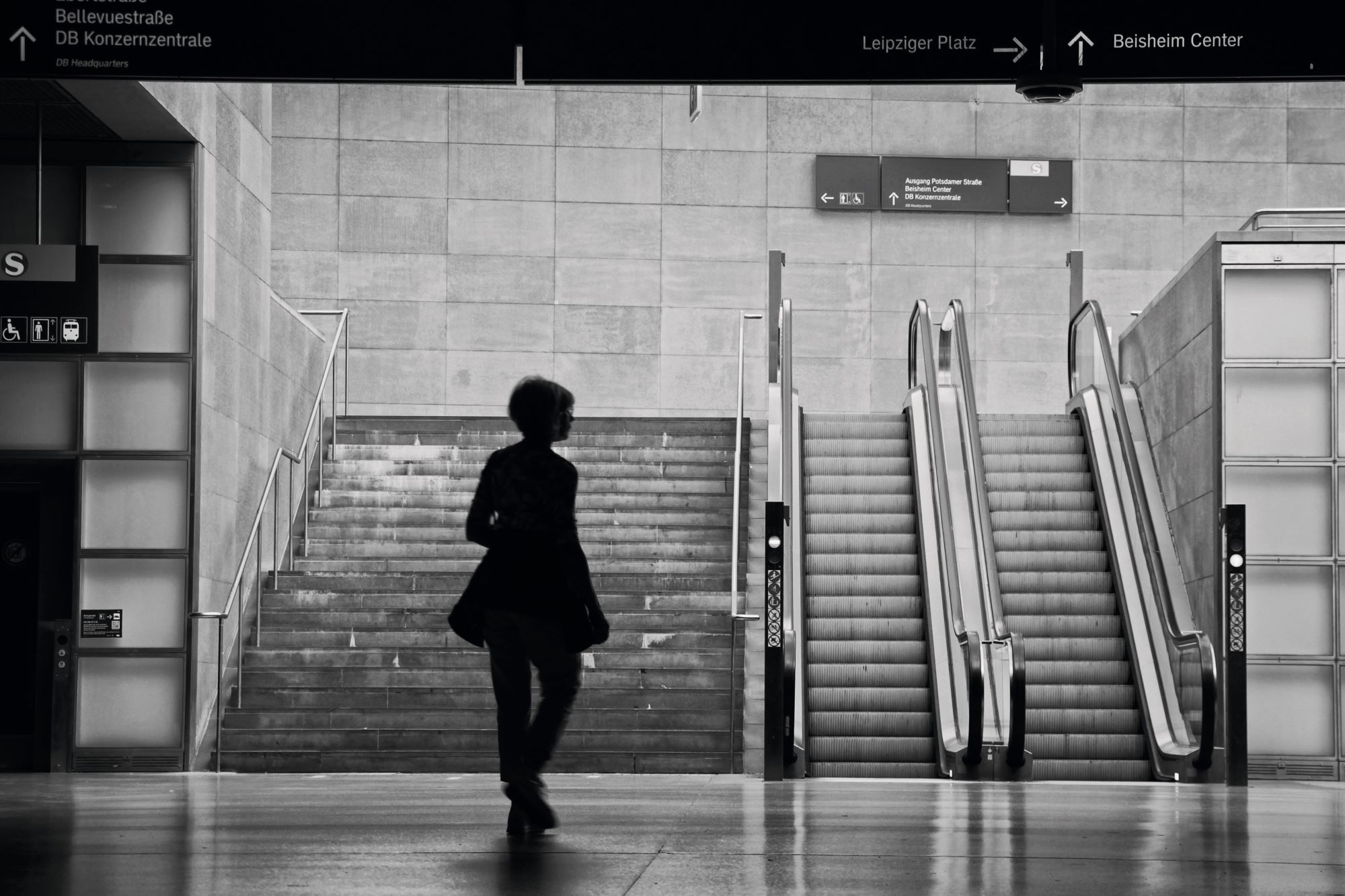 Eine Person geht auf eine Treppe und eine Rolltreppe in einer modernen überdachten Verkehrsstation zu; darüber hängen Schilder mit Richtungsangaben. (KI generierte Beschreibung)