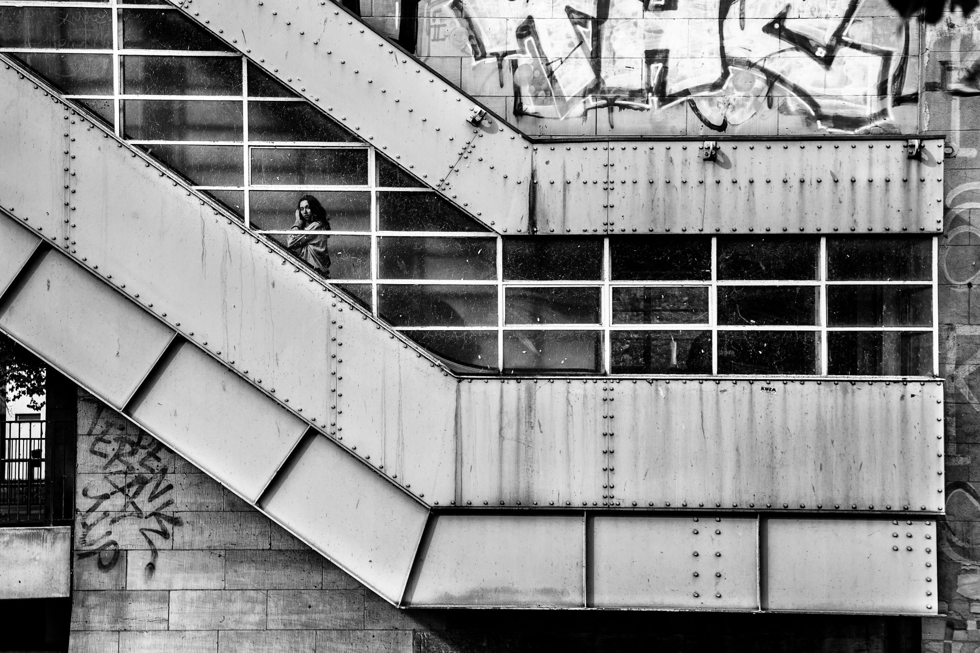 Eine Person geht eine geschlossene Außentreppe mit großen Fenstern und Graffiti an den umgebenden Beton- und Metallwänden hinunter. (KI generierte Beschreibung)