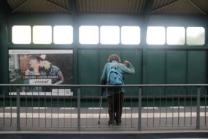 Eine Person mit einem blauen Rucksack und einem Gehstock steht auf einem Bahnsteig und wendet sich ab. An der Wand hinter ihnen ist eine Werbung zu sehen. (KI generierte Beschreibung)
