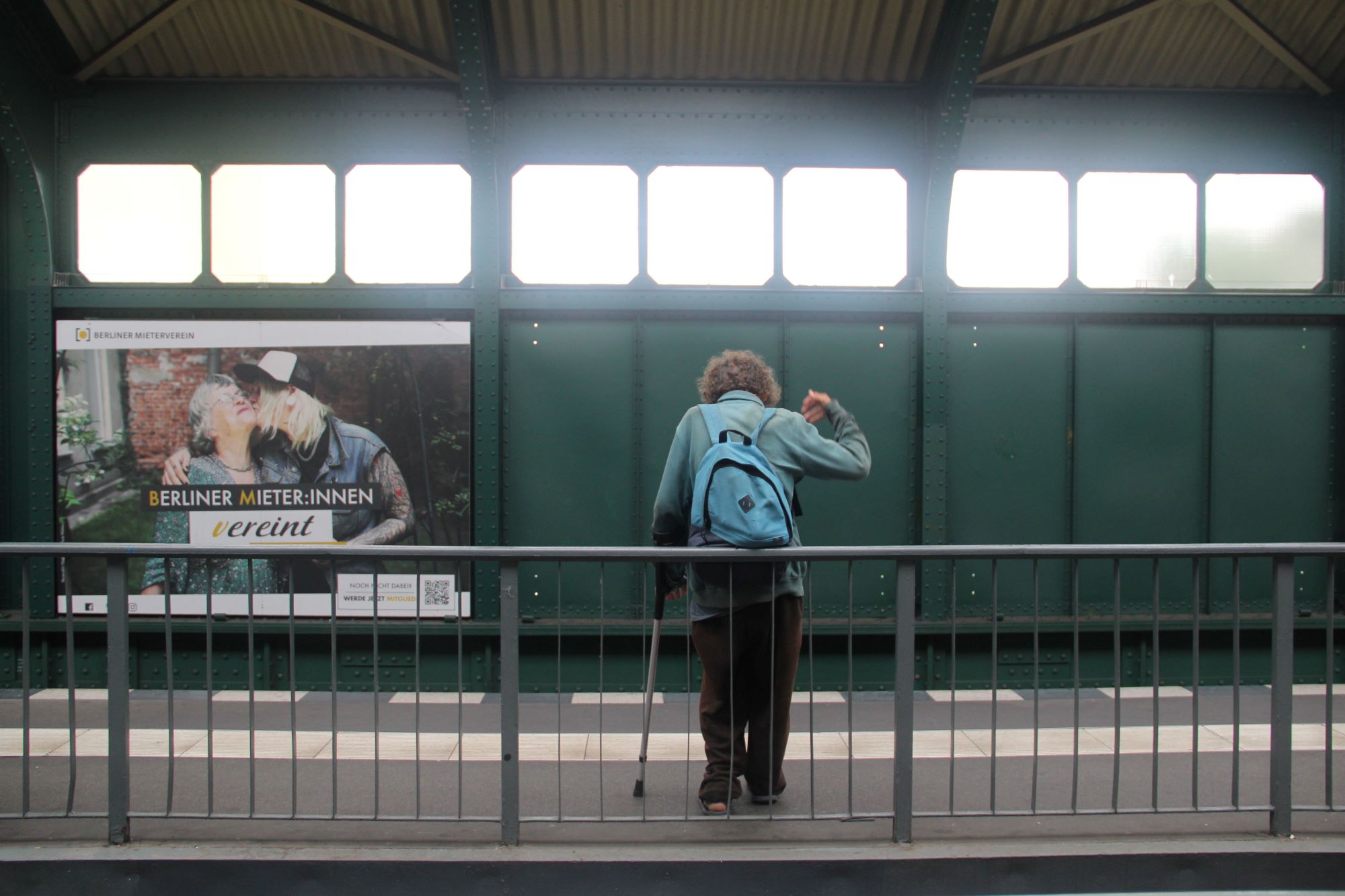 Eine Person mit einem blauen Rucksack und einem Gehstock steht auf einem Bahnsteig und wendet sich ab. An der Wand hinter ihnen ist eine Werbung zu sehen. (KI generierte Beschreibung)