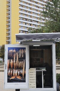 Vor einem hohen gelben Wohnhaus steht ein kleiner Straßenstand, an dem geräucherter Fisch verkauft wird; im Inneren des Standes hängen verschiedene Fische und ein Schild mit Preisangaben. (KI generierte Beschreibung)