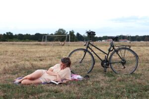 Eine Person liegt auf einer Decke und liest ein Buch auf einer Wiese neben einem schwarzen Fahrrad, mit einem Fußballtor und Bäumen im Hintergrund. (KI generierte Beschreibung)