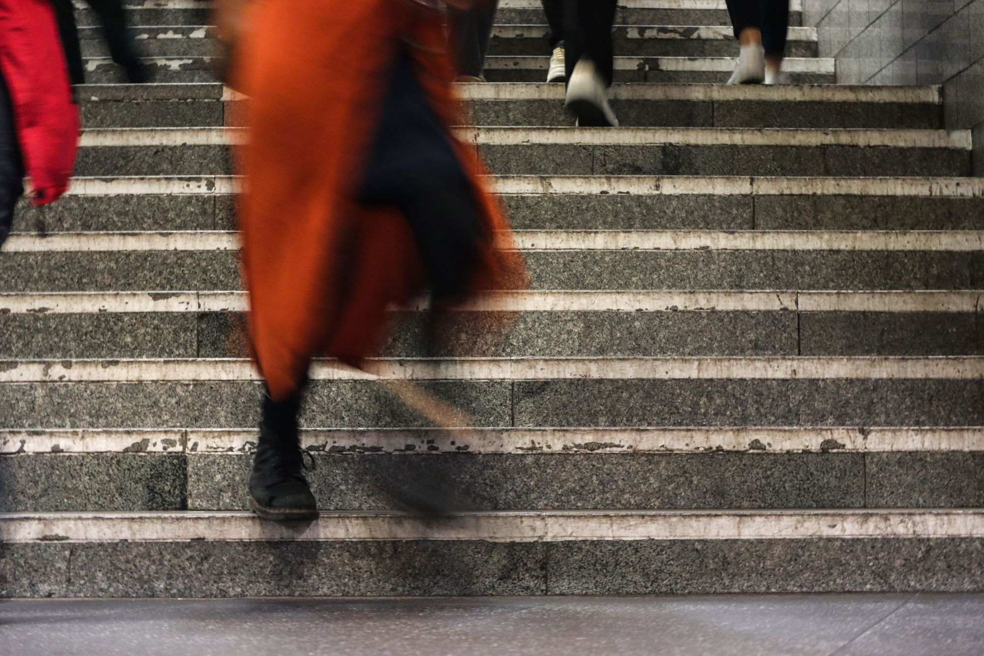 Unscharfe Gestalten gehen eine Steintreppe hinauf, wobei der Fokus auf einer Person in einem orangefarbenen Mantel und schwarzen Schuhen in Bewegung liegt. (KI generierte Beschreibung)
